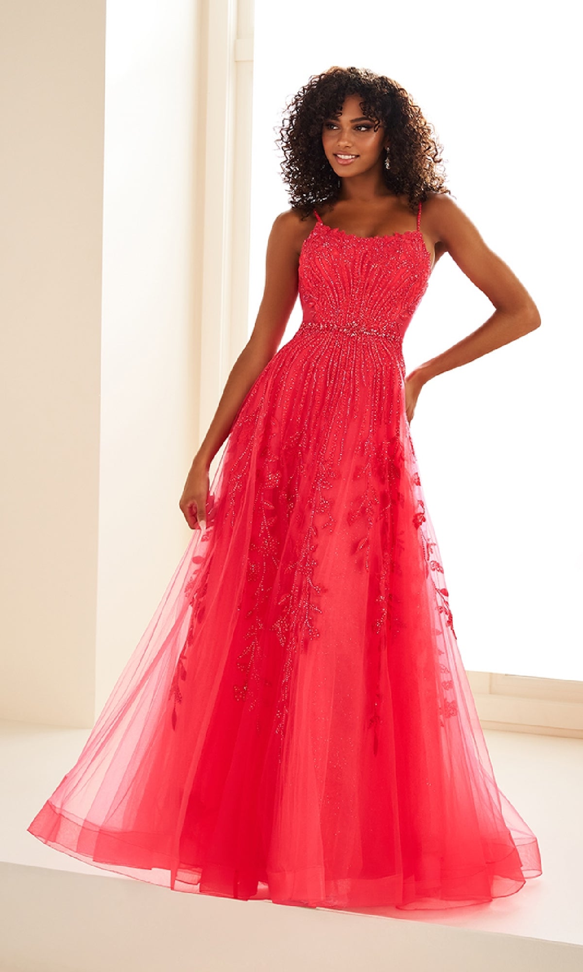 A woman in the Ellie Wilde EW36068, a sleeveless, floor-length bright pink prom dress with beaded details and glitter tulle, stands in a brightly lit room.